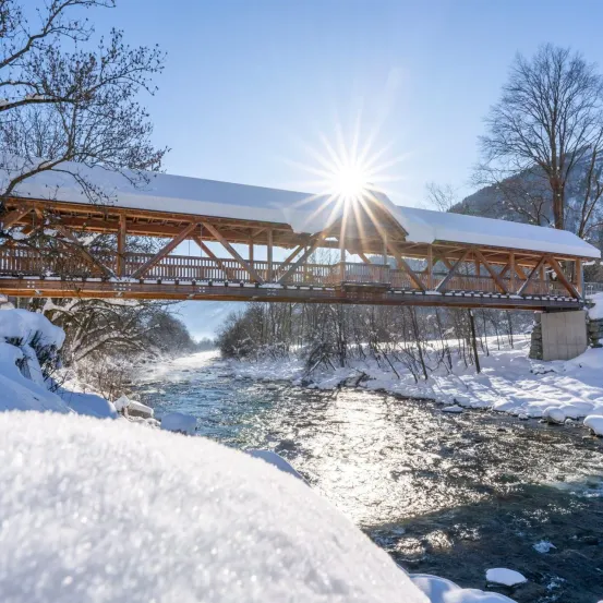 Eine Holzbrücke überspannt einen Fluss in einer verschneiten Landschaft. Schnee bedeckt die Brücke und die Umgebung. Die Sonne scheint hell darüber.