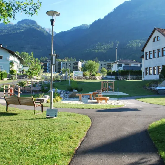 Eine friedliche Parkszene mit einem Weg, der zu einer Bank, einem Spielplatz und einem weißen Gebäude führt, alles unter einem klaren Himmel mit Bergen im Hintergrund.