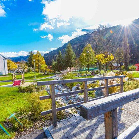 Ein sonniger Tag in einem Bergpark mit einer Holzterrasse, einem kleinen Wasserlauf und einem Spielplatz mit einer Rutsche unter einem blauen Himmel mit Wolken.