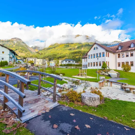 Eine Holzbrücke über einen kleinen Bach führt zu einem Park mit einem Spielplatz. Hinter dem Park stehen Häuser und ein Gebäude mit Solarpanelen. In der Ferne befinden sich Berge.