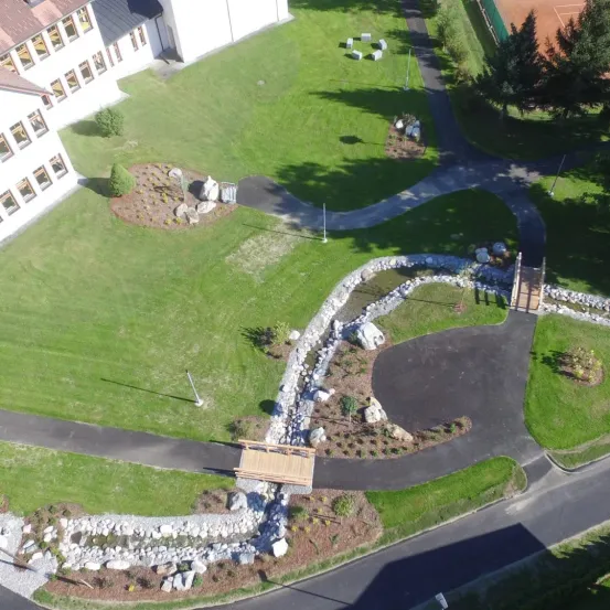 Luftaufnahme eines großen Gebäudes mit mehreren Fenstern, umgeben von grünem Rasen und einem gepflasterten Weg. Bäume und Pflanzen sind im gesamten Bereich verstreut.