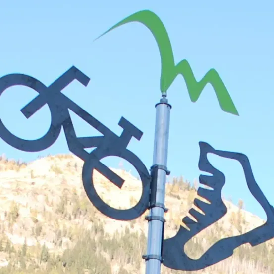 Ein Metallschild mit Fahrrad- und Schuhdesign vor einer Kulisse aus Berg, Bäumen und klarem Himmel.