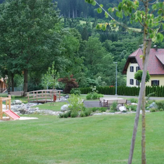 Ein Spielplatz mit einem roten Rutschen und einer Holzbrücke, umgeben von Bäumen und Pflanzen. Ein Haus mit rotem Dach ist in der Ferne.