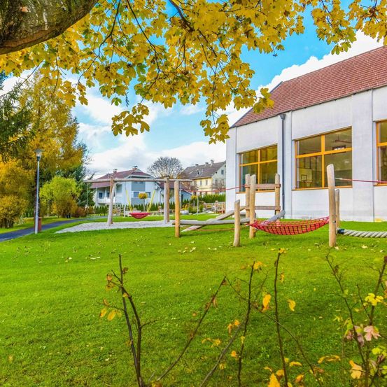 Ein Spielplatz mit einem roten Hängematten und Klettergerüste in einer sonnigen, grünen Parkfläche. Der Spielplatz ist von einem Zaun umgeben, mit Bäumen und Pflanzen in der Nähe. Ein Gebäude mit Glasfenstern und einem braunen Dach ist im Hintergrund sichtbar.