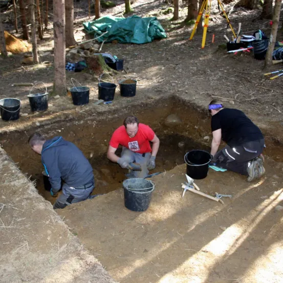 Drei Personen graben in einer Grube in einem Waldgebiet. Einer trägt ein rotes Shirt, ein anderer eine blaue Kapuzenjacke und der dritte ein schwarzes Shirt. Sie haben Eimer und Werkzeuge um sich herum.