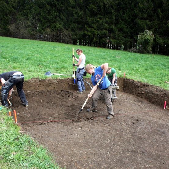 Eine Gruppe von Menschen ist in einem Grasfeld mit einer Ausgrabung beschäftigt, wobei zwei Personen Schaufeln benutzen und eine andere einen Schubkarren schiebt.