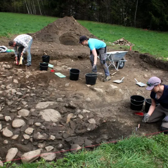 Drei Personen sind mit Ausgrabungsarbeiten beschäftigt. Ein Mann benutzt eine Schaufel, ein weiterer kniet mit einer Bürste und ein Dritter schiebt einen Schubkarren. Mehrere Eimer sind um die Stelle herum platziert.