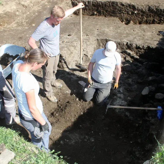 Drei Personen arbeiten auf einer Baustelle, eine kniet in einem Loch und zwei andere stehen mit Schaufeln. Das Loch ist mit Erde und Steinen gefüllt.