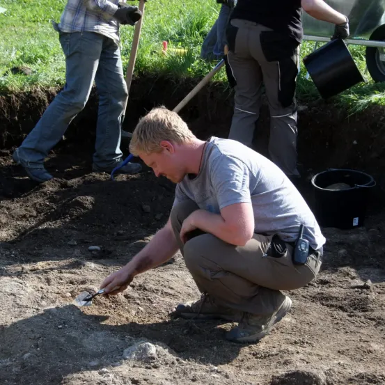 Ein Mann kniet in der Erde und benutzt eine Schaufel zum Graben. Hinter ihm stehen zwei Personen, eine hält eine Schaufel. Sie befinden sich in der Nähe eines Schubkarrens und Eimer.