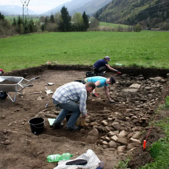 Archäologen graben in einem Feld mit Steinen, einem Schubkarren und einem klaren blauen Himmel darüber. Eine Person beugt sich mit einem Pinsel vor.