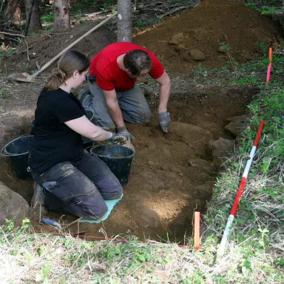 Zwei Personen graben in einem Loch, tragen Handschuhe und Stiefel. Eine Frau kniet mit einem Eimer, während ein Mann über dem Boden hockt. Eine Schaufel liegt in der Nähe auf dem Boden.