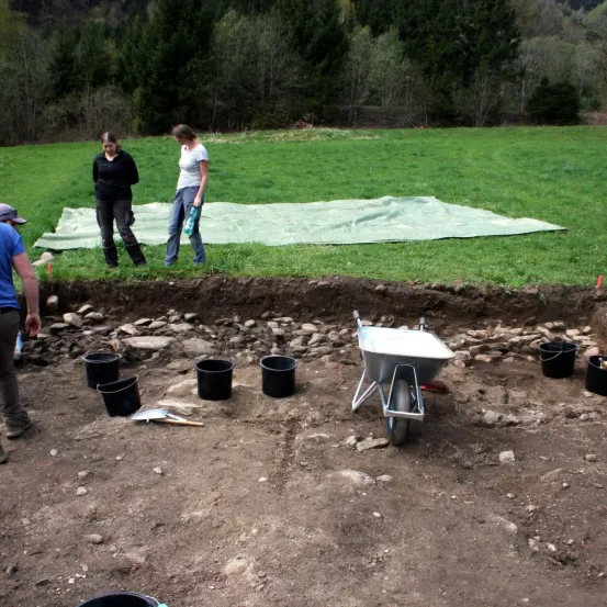 Drei Personen stehen in einem Feld mit einer archäologischen Ausgrabung. Ein Schubkarren und mehrere Eimer sind in der Nähe. Das Gebiet ist mit einer grünen Plane bedeckt.