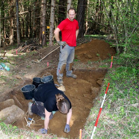 Zwei Personen im Wald graben mit Eimern und Werkzeugen in den Boden, einer steht und beobachtet, während der andere kniet und gräbt.
