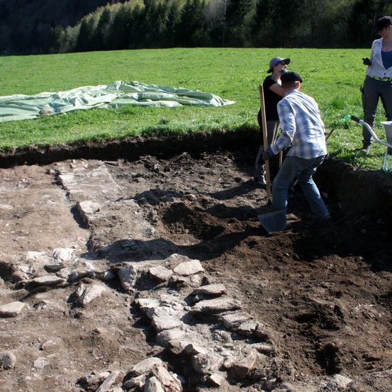 Drei Personen graben in einem Feld mit Steinen. Eine Person benutzt eine Schaufel, während eine andere einen Holzstab hält. Eine Frau mit einer Schubkarre steht in der Nähe.
