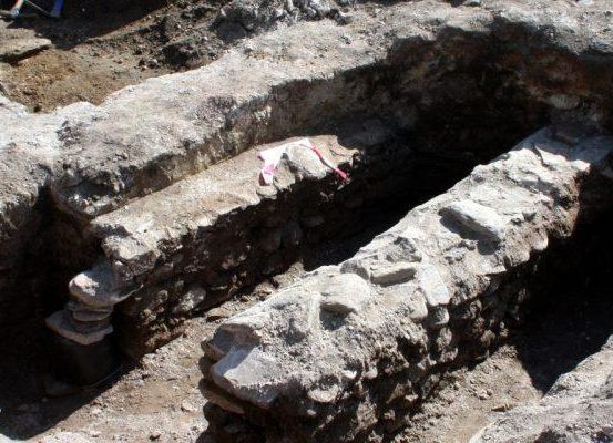 Ein Ausgrabungsbereich mit einer großen Steinstruktur, die eine rechteckige Öffnung mit einer Steinmauer und einer rosa Markierung darauf zeigt.