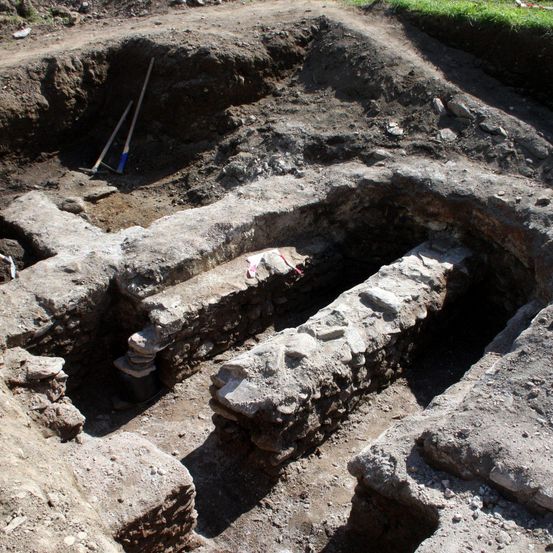 Eine archäologische Ausgrabungsstätte mit Steinstrukturen, einschließlich einer Steinbank, umgeben von Erde und Steinen. Werkzeuge sind im Boden platziert.