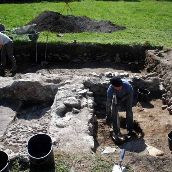 Archäologen arbeiten an einer Ausgrabungsstätte, einer gräbt und der andere schiebt einen Schubkarren. In der Nähe befinden sich ein Haufen Erde, Werkzeuge und Eimer.