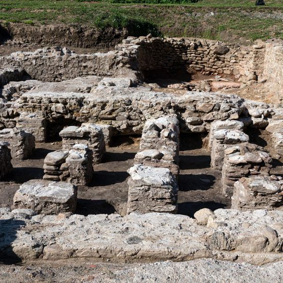 Ein Blick von oben auf eine archäologische Ausgrabungsstätte mit Steinstrukturen, von denen einige teilweise eingestürzt sind. Der Boden ist steinig, und Schatten, die von den Steinen geworfen werden, sind sichtbar. Im Hintergrund deutet ein Grasbereich auf eine ländliche Umgebung hin.