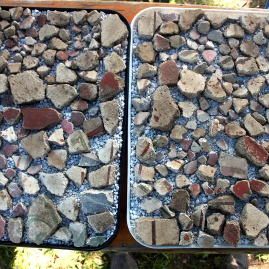 Zwei Schalen mit gemischten Steinen und Kieselsteinen, einschließlich zerbrochener Ziegelbruchstücke, liegen auf einer Holzoberfläche. Die Steine variieren in Farbe und Größe, wobei einige rote und weiße Streifen aufweisen.