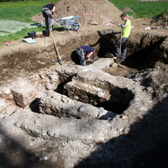 Drei Arbeiter sind an einem archäologischen Ausgrabungsstandort. Einer steht mit einem Schubkarren, einer kniet auf dem Boden und ein anderer steht mit einer Schaufel. Der Standort ist von Erde und Felsen umgeben.