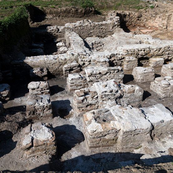 Eine Luftaufnahme einer Steinruinenstätte, teilweise ausgegraben, mit verstreuten Steinen und Überresten von Mauern, die Schatten auf dem Boden werfen.