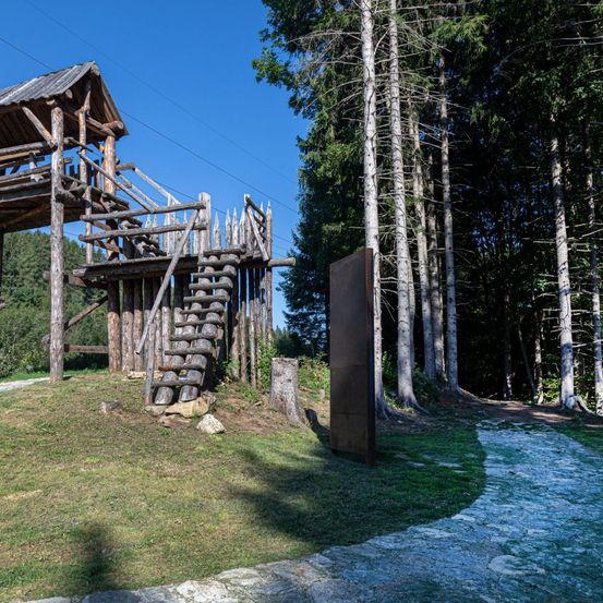 Eine Holzstruktur mit Treppen und Dach steht neben einem Metallobjekt und ist von einem Wald mit einem Steinweg umgeben.
