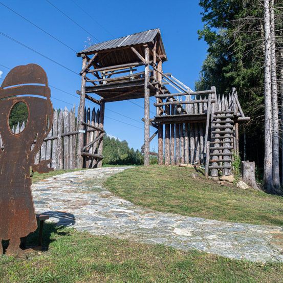 Ein verwittertes Holzgebäude steht mit einem Pfad, der zu einem Tor unter einem klaren blauen Himmel führt. In der Nähe ist eine rostige Skulptur einer Figur auf einem Rasenfeld platziert.