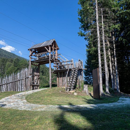Ein hölzerner Wachturm und eine Treppe stehen auf einem Grashügel, umgeben von einem Holzzaun und hohen Bäumen. Der Weg führt zum Turm, mit einem klaren Blick auf die Berge in der Ferne.