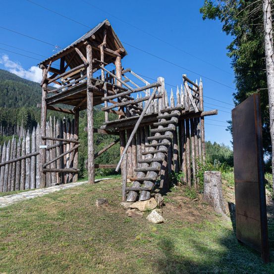 Ein hölzerner Wachturm mit einer Treppe und einem Zaun steht auf einem Hügel unter einem klaren blauen Himmel.