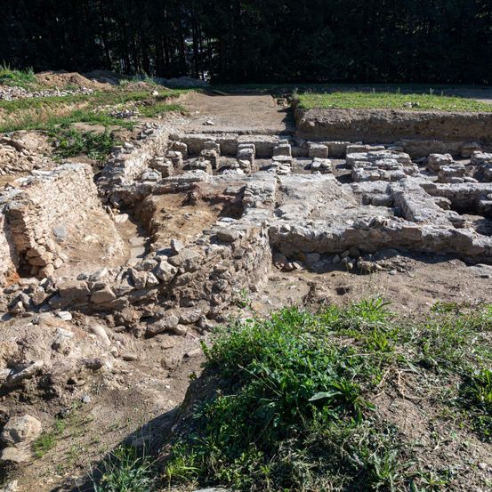 Archäologische Ausgrabungsstätte mit Steinfundamenten, umgeben von Erde und Gras. Bäume im Hintergrund.
