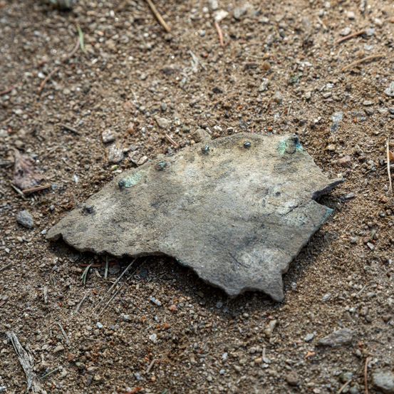 Ein Metallfragment mit rauher Oberfläche, möglicherweise von einem alten Artefakt, liegt auf sandigem Boden mit sichtbarem Schmutz und kleinen Steinen.