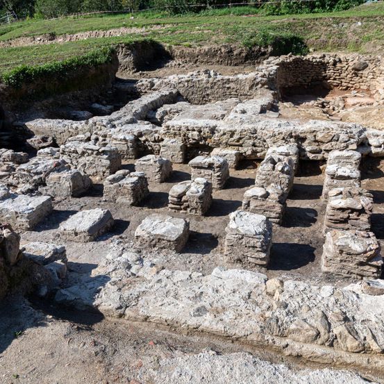 Ein archäologischer Fundplatz mit sichtbaren Steinstrukturen, möglicherweise Überreste eines alten Gebäudes. Das Gebiet ist von einem Grasfeld umgeben und zeigt Spuren einer Ausgrabung.