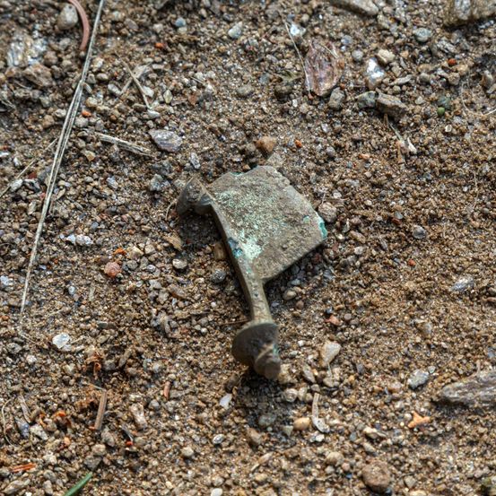 Ein altes, korrodiertes Metall-Beil liegt auf dem Boden, teilweise in Erde vergraben und umgeben von kleinen Steinen und Schutt.