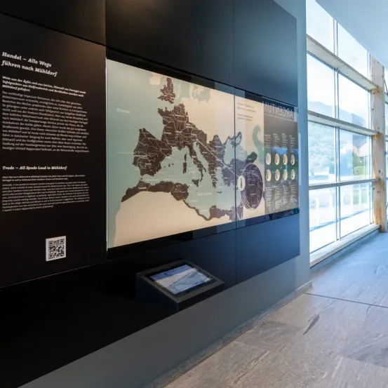 Eine große Karte und ein Tablet-Display befinden sich an einer Wand, mit Text darüber und darunter. Der Text lautet 'Alle Wege führen nach Mühldorf'. Ein QR-Code befindet sich links neben der Karte. Der Raum hat große Fenster.