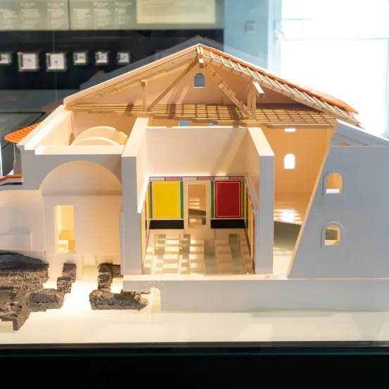 Ein Modell eines alten Gebäudes mit farbenfrohen Wandmalereien, das unter Glas ausgestellt ist. Die Struktur hat ein Ziegeldach, gewölbte Türen und einen teilweise geöffneten Eingang.