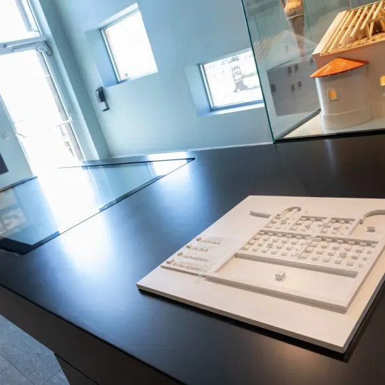 Eine moderne Ausstellung mit schwarzem Arbeitstisch zeigt ein weißes architektonisches Modell. Dahinter zeigt ein Glasvitrine ein Miniaturhaus mit gefliestem Dach. Der Raum hat weiße Wände und Fenster.