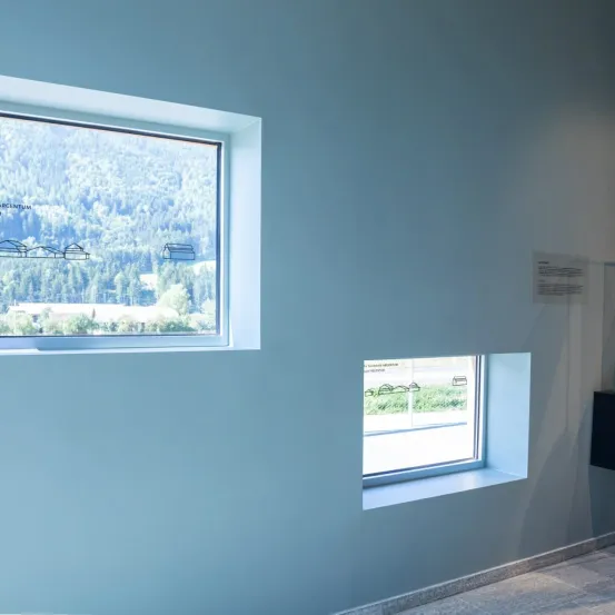 Ein Innenbereich mit einem großen Fenster auf der linken Seite und einem kleineren Fenster auf der rechten Seite. Beide Fenster bieten panoramische Ausblicke auf die umliegenden Berge.