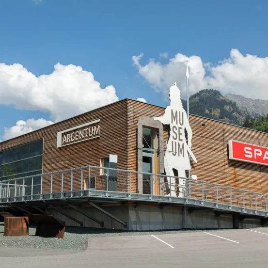 Ein modernes Gebäude mit Holzfassaden und einem Balkon zeigt eine große weiße Silhouette eines Ritters. Es heißt Argentum. Im Hintergrund sind Berge zu sehen.