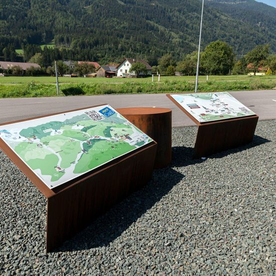 Zwei Holzbänke mit Karten stehen auf Kies am Straßenrand, mit Bergen und Häusern im Hintergrund.