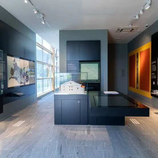 Ein Innenraum eines Museums mit einem kleinen architektonischen Modell unter einer Glasvitrine, einer großen Karte an der Wand und grauen Bodenfliesen.