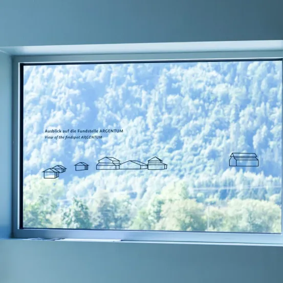Ein Fensterausblick zeigt eine Landschaft mit Bergen und Bäumen. Es gibt eine Beschriftung auf Deutsch, die 'Ausblick auf die Fundstelle ARGENTUM' lautet. Es sind einfache Illustrationen von Gebäuden zu sehen.