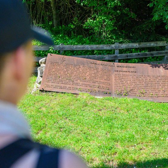 Eine Person steht auf Gras und betrachtet eine Gedenktafel mit Text, im Freien mit einem Holzzaun im Hintergrund.