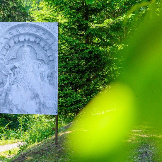 Eine Steinschnitzerei einer sitzenden Figur, möglicherweise eines Königs, mit kunstvollen Mustern ziert ein Schild im Wald. Die Schnitzerei wird von Sonnenlicht beleuchtet, das Schatten auf die nahen Bäume und den Boden wirft.