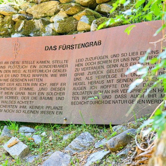 Eine braune Plakette mit Text darauf befindet sich in einem Grasbereich. Die Plakette zeigt eine historische Erzählung über das Grab eines Fürsten.