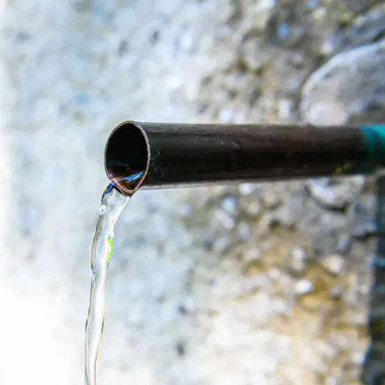 Eine Nahaufnahme einer Röhre, aus der Wasser in einem gleichmäßigen Strom fließt. Die Röhre ist metallisch mit einer grünlich-blauen Oberfläche.