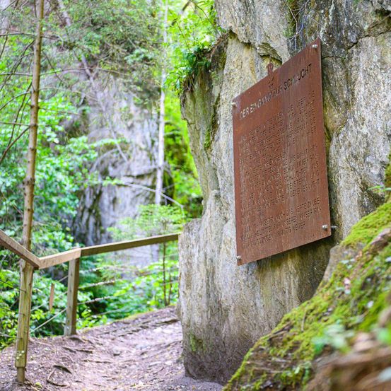 Eine bronzene Plakette ist an einer Steinwand befestigt, möglicherweise in einem Wald. Links befindet sich ein hölzerne Geländer und rechts ein moosbewachsener Felsen.