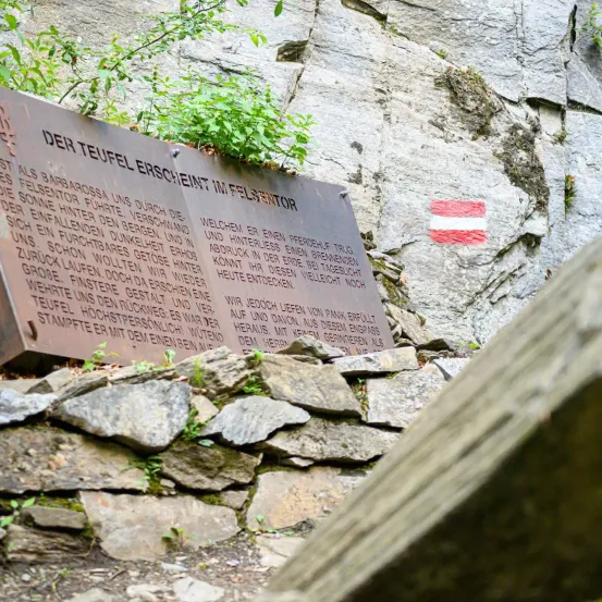 Eine Plakette mit deutschem Text ist an einer felsigen Wand montiert, teilweise von Steinen und Moos bedeckt. Darüber befindet sich ein rot-weißes Zeichen.