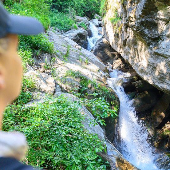 Eine Person mit einer Mütze beobachtet einen Wasserfall, der in einem üppigen, grünen Tal über felsige Klippen strömt.