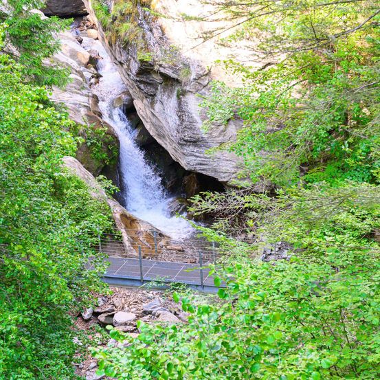 Ein Wasserfall stürzt durch einen felsigen Berg in einem üppig grünen Wald, mit einer Metallbrücke, die das Wasser überspannt.