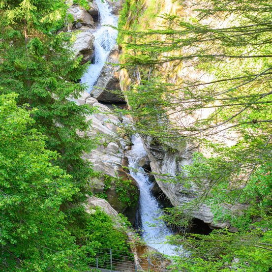 Ein Wasserfall stürzt an von üppigem grünem Laub umgebenen Felsen hinab, mit einem Metallgeländer in der Nähe.